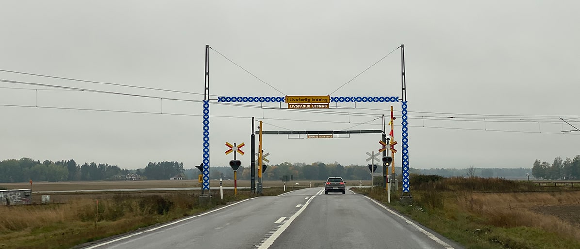 En blå och vit järnvägsövergångsgrind syns på höger sida av en tom motorväg i Sverige, med två gula skyltar på varje ände. En bil syns svagt vid ett hörn nära järnvägsspåren. Bakgrunden visar ett platt landskap och grå himmel. 